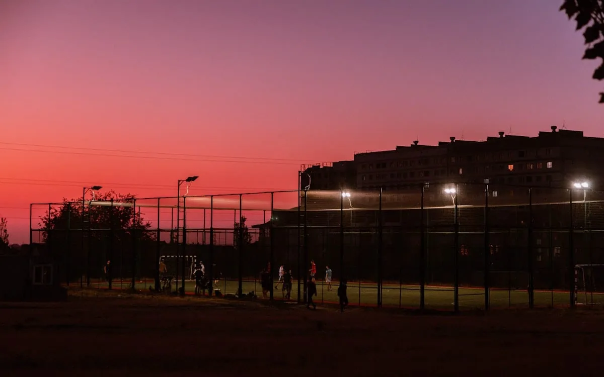 Floodlit public sports courts in front of a tall apartment block at dusk, people still playing as the sky turns pink