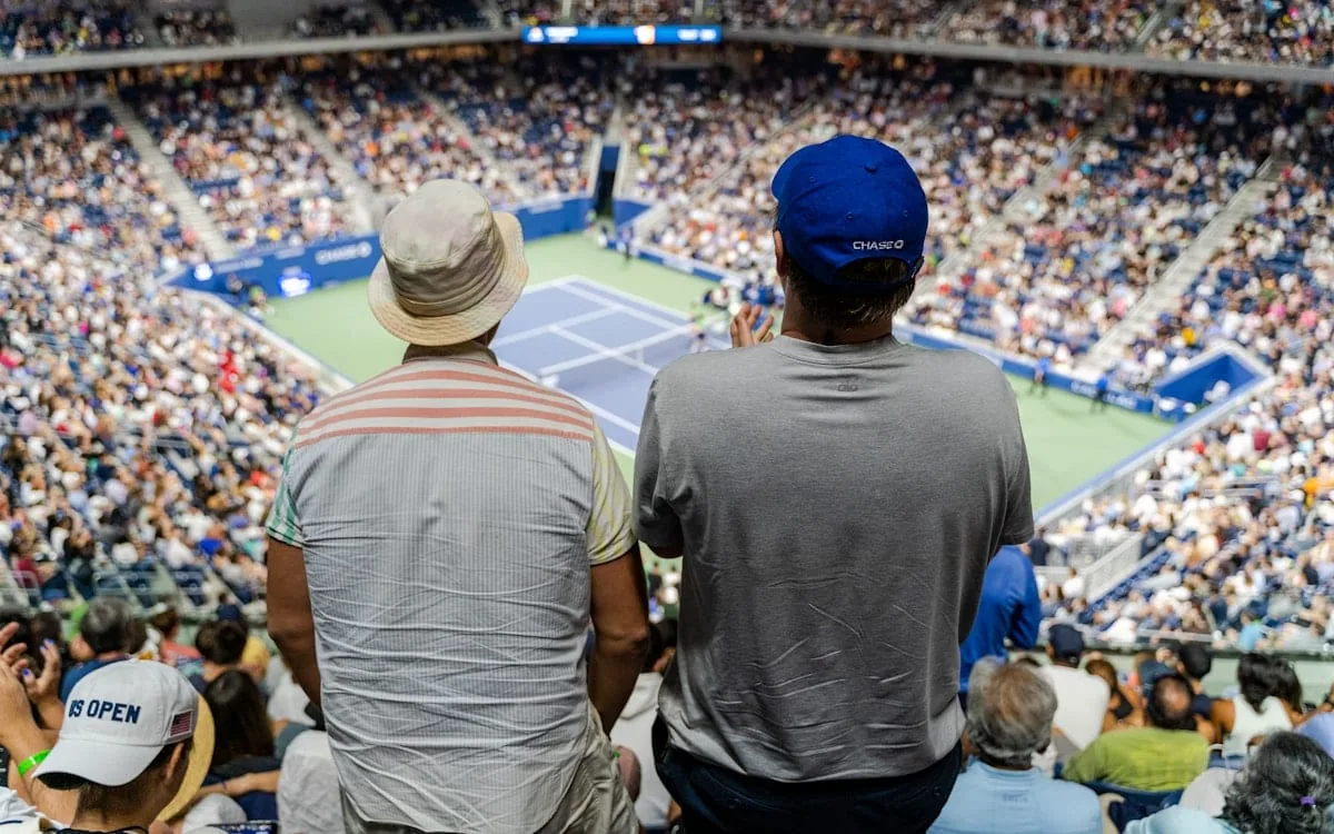 A packed tennis stadium seen from the upper rows, two fans standing shoulder to shoulder watching the court far below